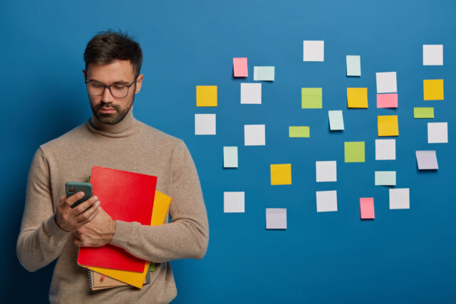 Indoor view of bearded man uses mobile phone for online conversation, holds textbook, wears spectacles, dressed in glasses and brown sweater, looks for information, colorful notes behind on wall